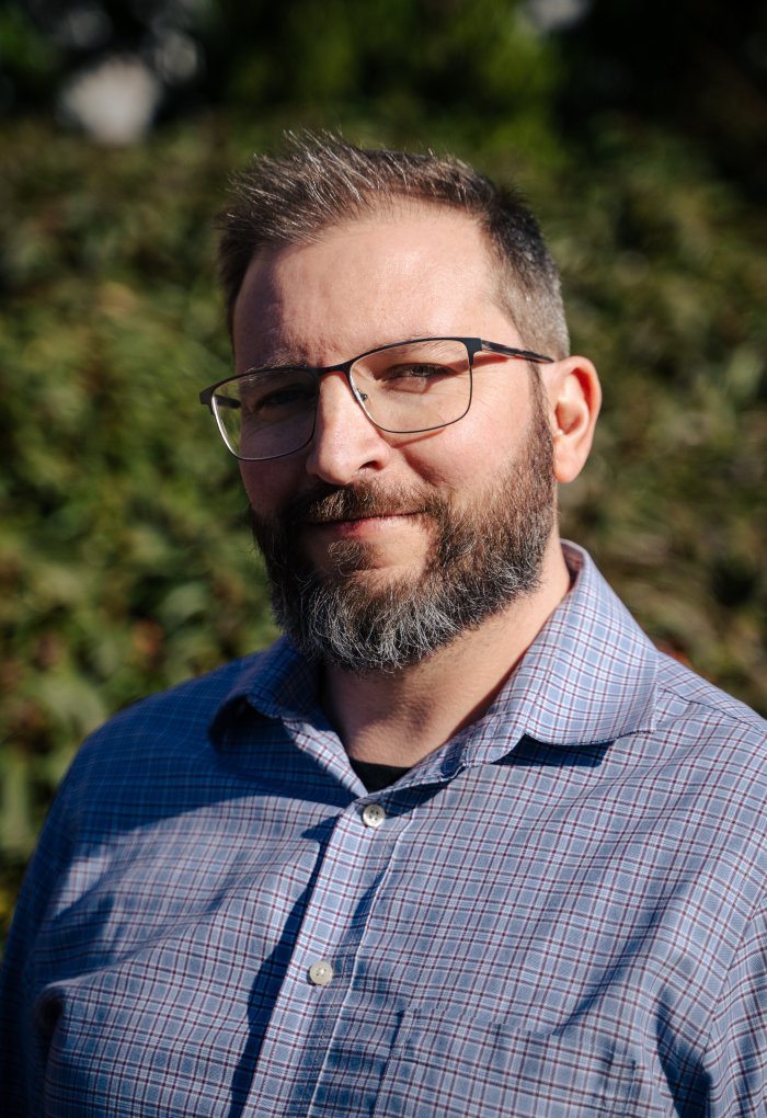 A man in a blue plaid shirt with glasses and a beard poses for a headshot.