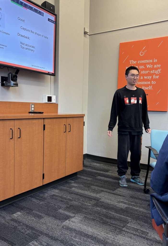 A man in sweat pants and a black sweatshirt stands at the front of a room near a computer screen.