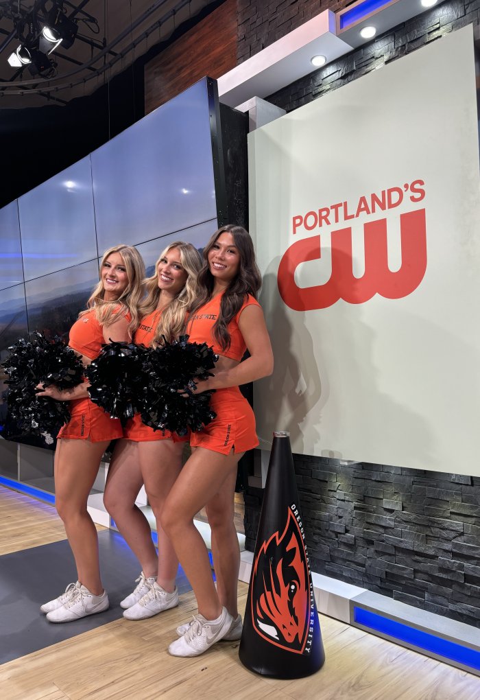 Three woman in orange OSU cheer uniforms stand beside each other, holding pompoms and smiling at the camera in a Portland news studio. 