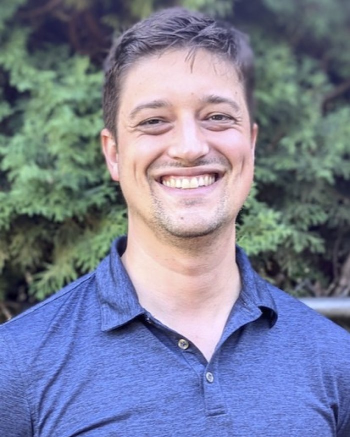 A man in a blue shirt smiling with trees in the background.
