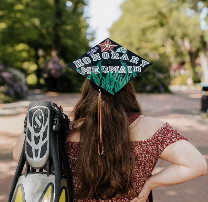 Simone Burton facing away from the camera, holding her scuba fins and wearing her decorated grad cap that says "Honorary Mermaid".
