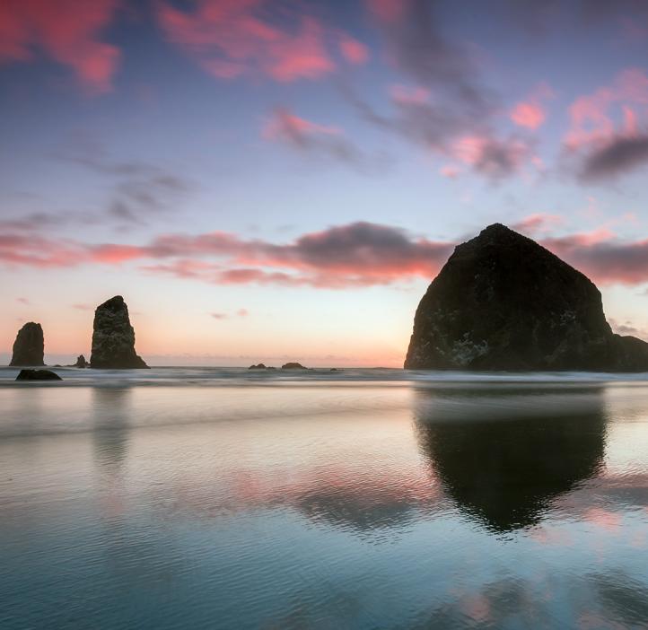 Three rocks jut up from Cannon Beach on the Oregon Coast during a sunset with pink clouds. 