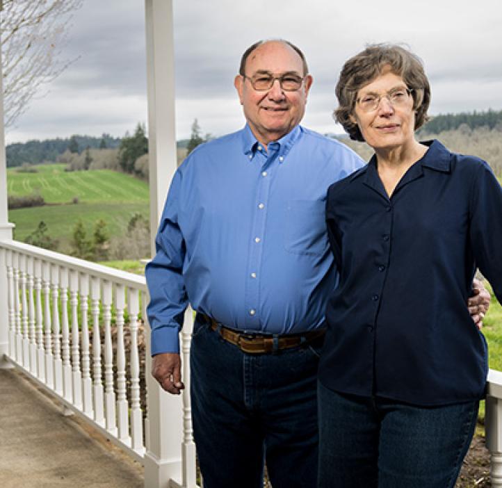 Norbert and Eileen Hartmann lean against a porch railing with green landscape behind them. 