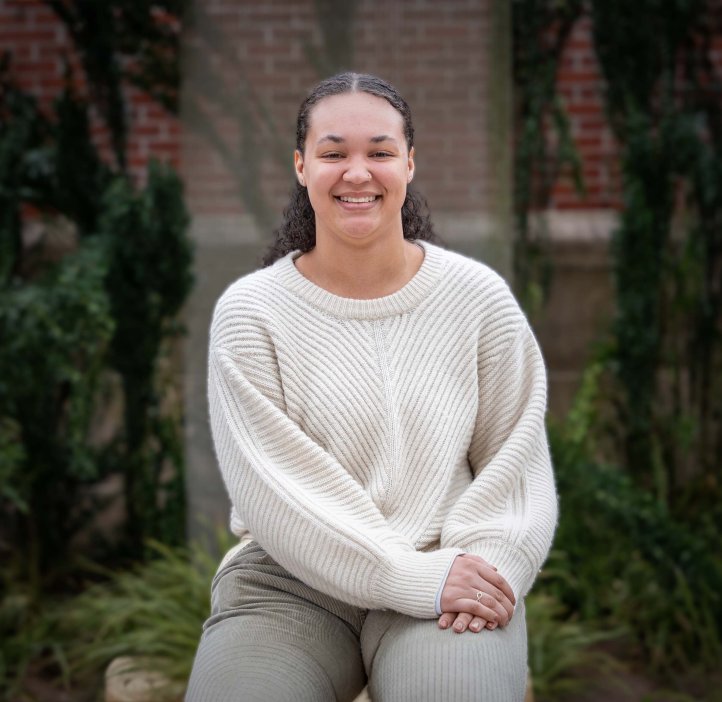 Camrie Smith sits with her hands in her lap wearing a white sweater. She is outdoors with a wall of vines behind her. Her natural hair is pulled back and she smiles broadly at the camera. 