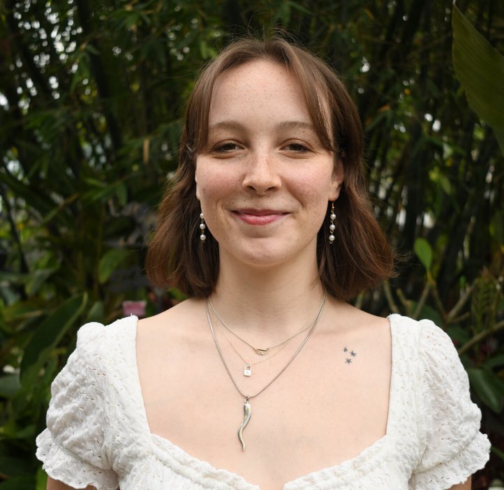 A woman in a white blouse stands in front of greenery. 