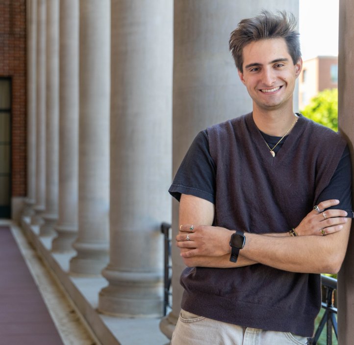 Physics student Diego Menendez leans on a large white column at Memorial Union Hall at Oregon State University. He has his arms crossed in front of him and he smiles at the camera. The hall behind him stretches on with a series of large white columns. 