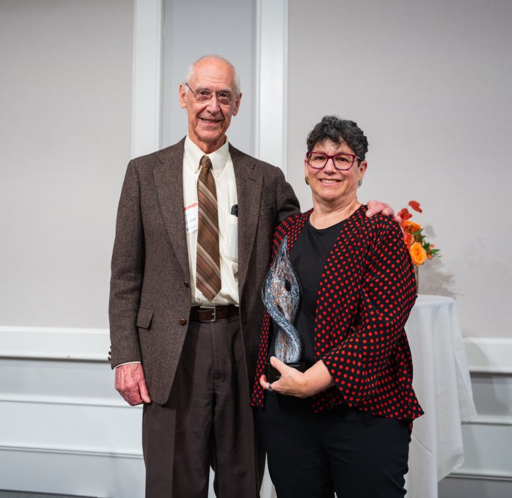 A man in a brown suit stands next to a woman wearing a red and black dress jacket. The woman is holing a glass award.