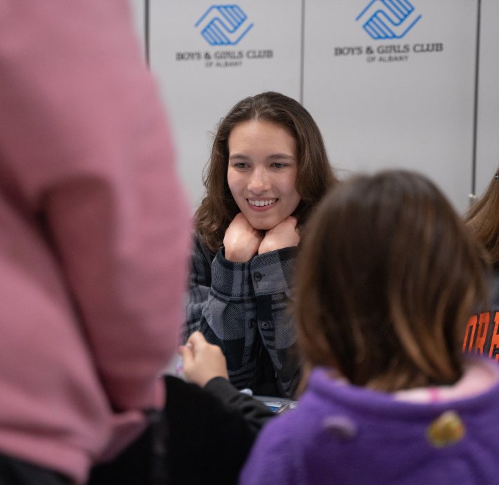 An OSU student has their hands tucked under their chin and their elbows on the table. She is smiling widely on the children across the table from her.