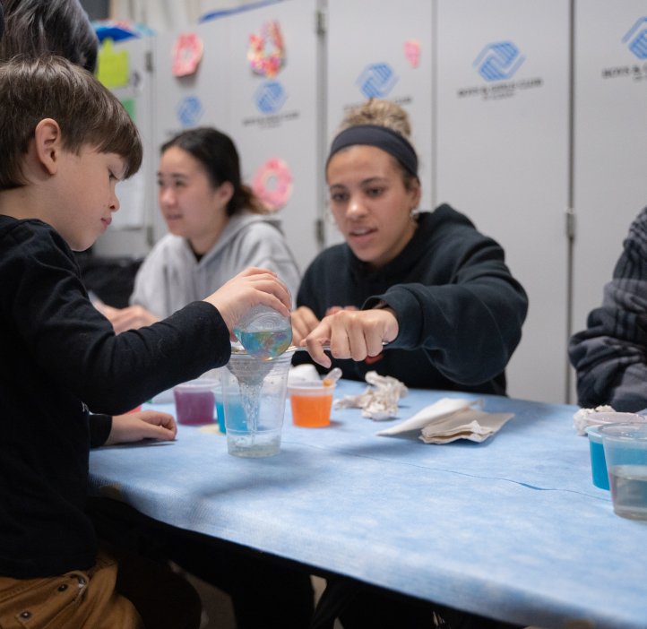 Three OSU students at a table with chromography experiments on the table in front of them. The one in the center is helping a child pour colorful liquid from a pitcher, into a cup. The student next to her has her mouth open in a wide smile, looking shocked. 