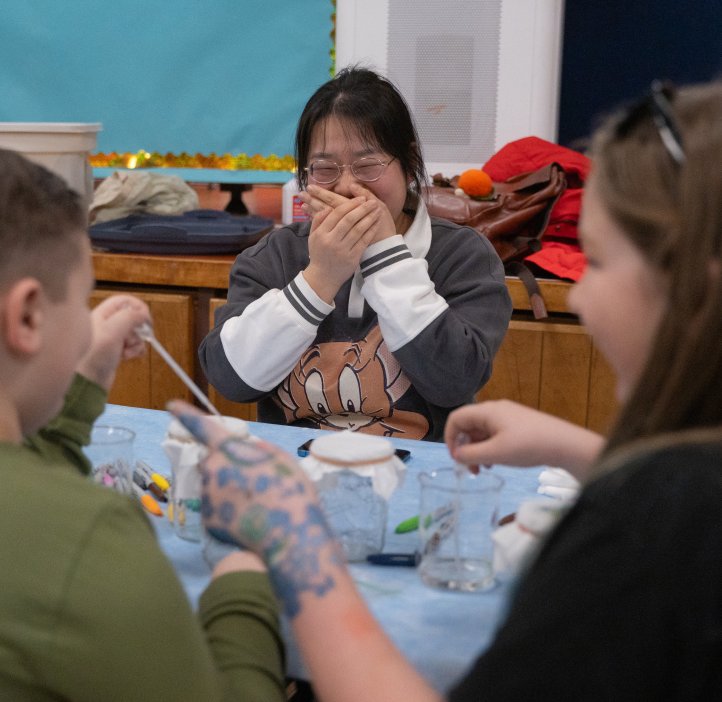 An OSU student covers her mouth while she laughs hard. There are two children in front of her engaged in a chromography experiment.