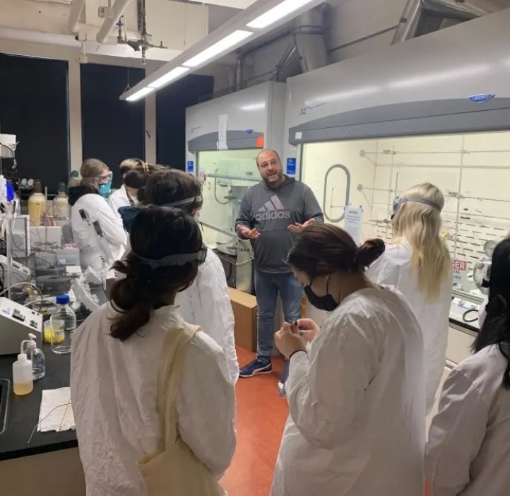 A group of students in lab coats stand around a professor in a laboratory.