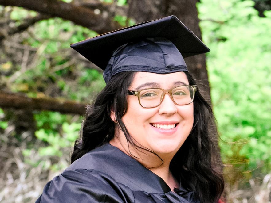 Angelica Ramirez standing in graduation gown in forest